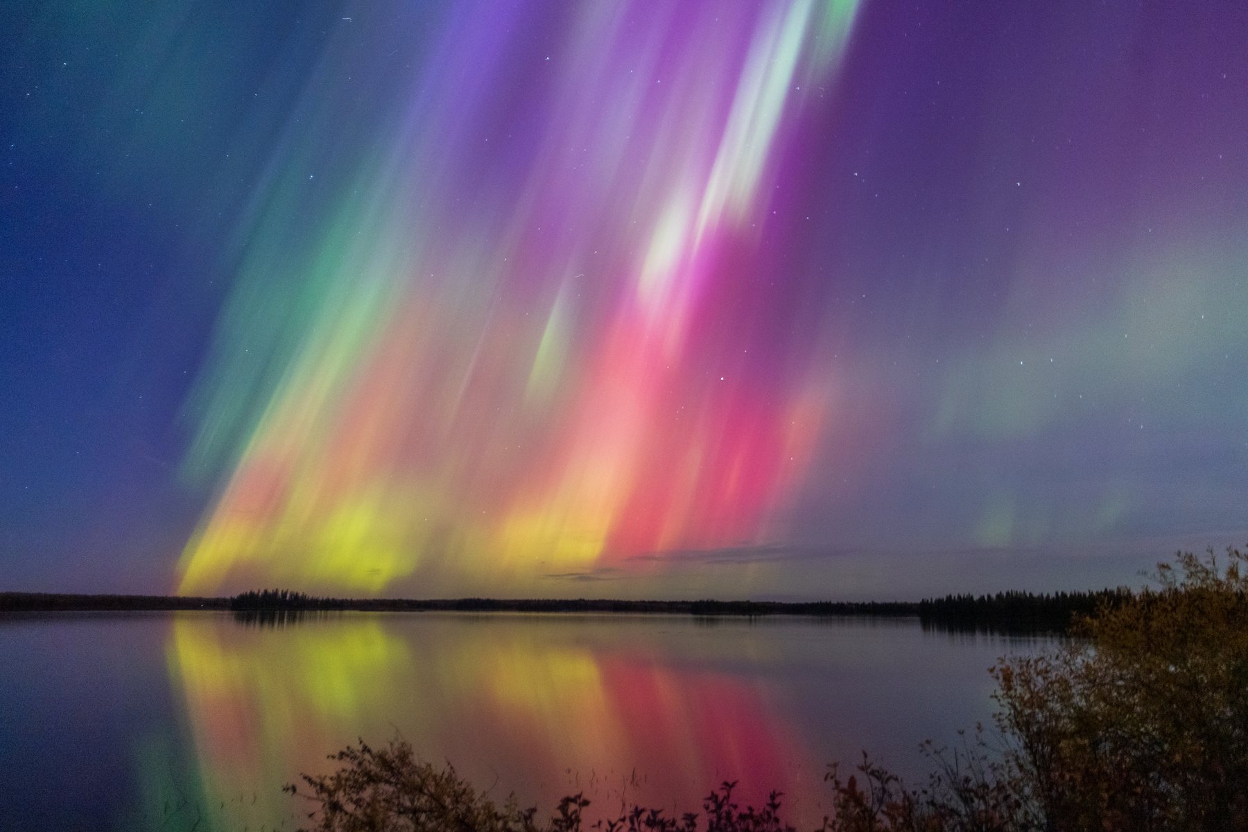Pink and green aurora curtains above the treeline, reflected in the lake