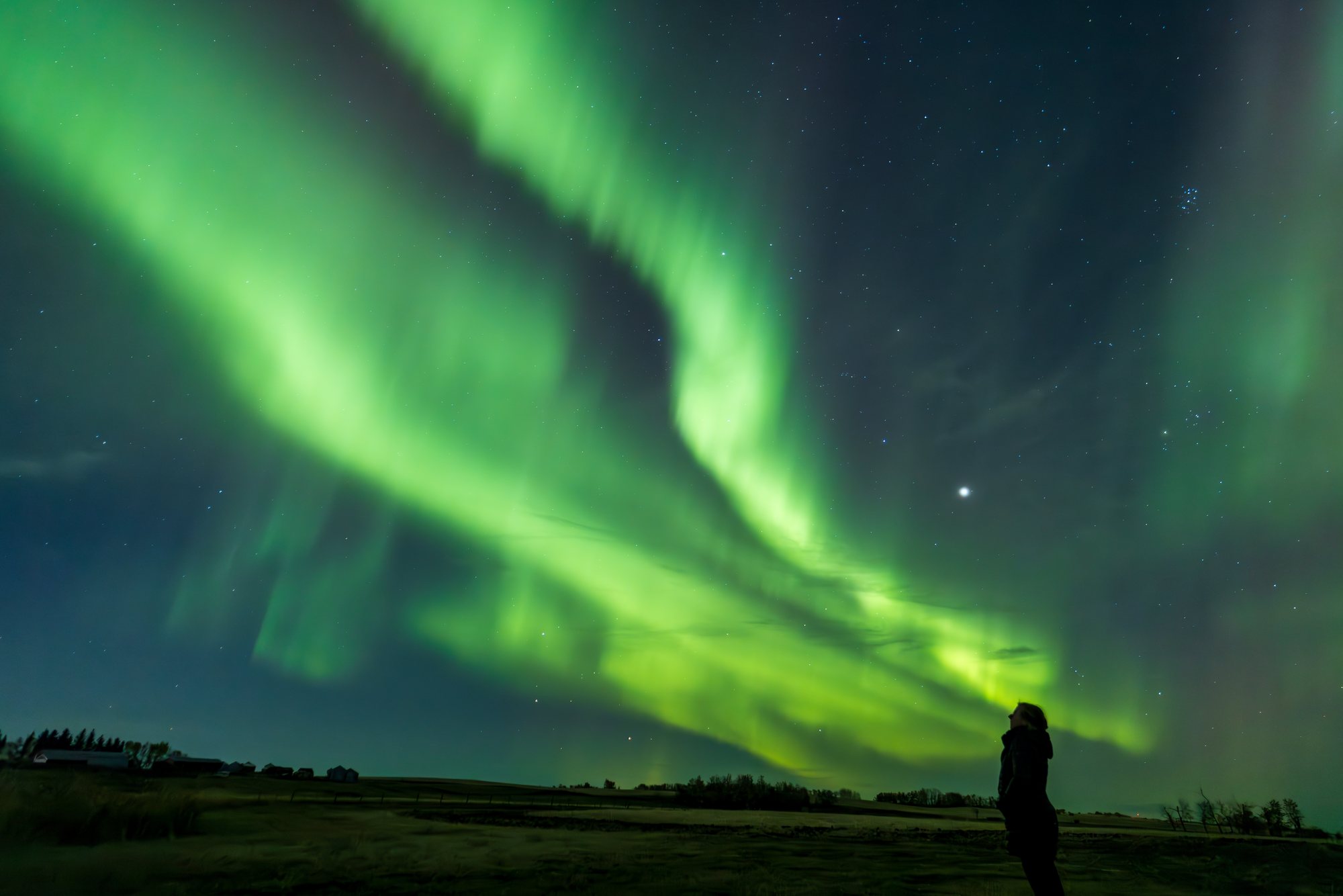 A lone figure silhouetted beneath a great arc of green aurora