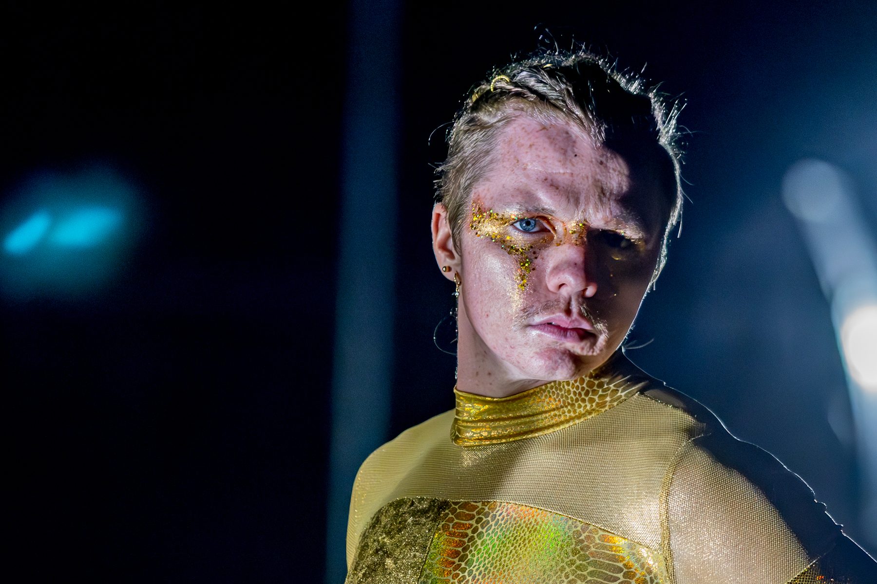A performer in gold with glitter face paint under stage light